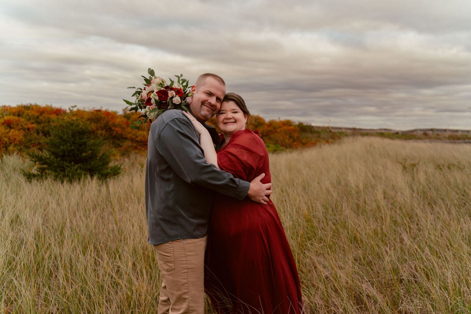 A couple embraces in a field of tall grass during autumn with cloudy skies overhead.