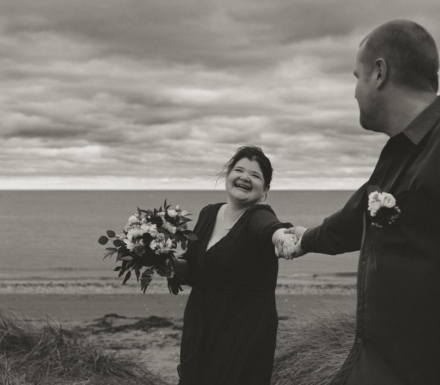 A joyful couple shares a candid moment on a beach with dramatic cloudy skies and ocean waves in the background.