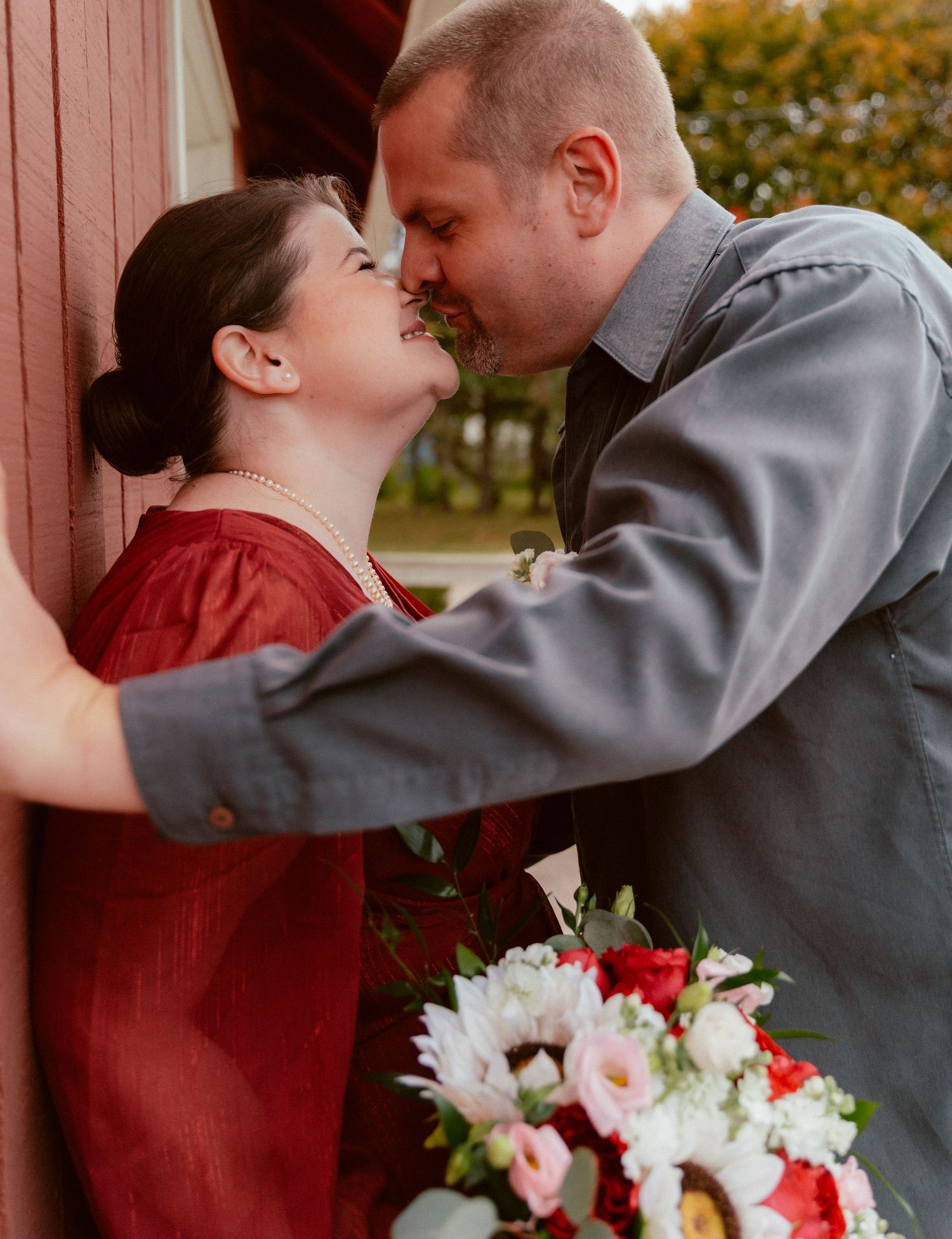 Romantic couple sharing intimate moment against red barn wall with floral bouquet.
