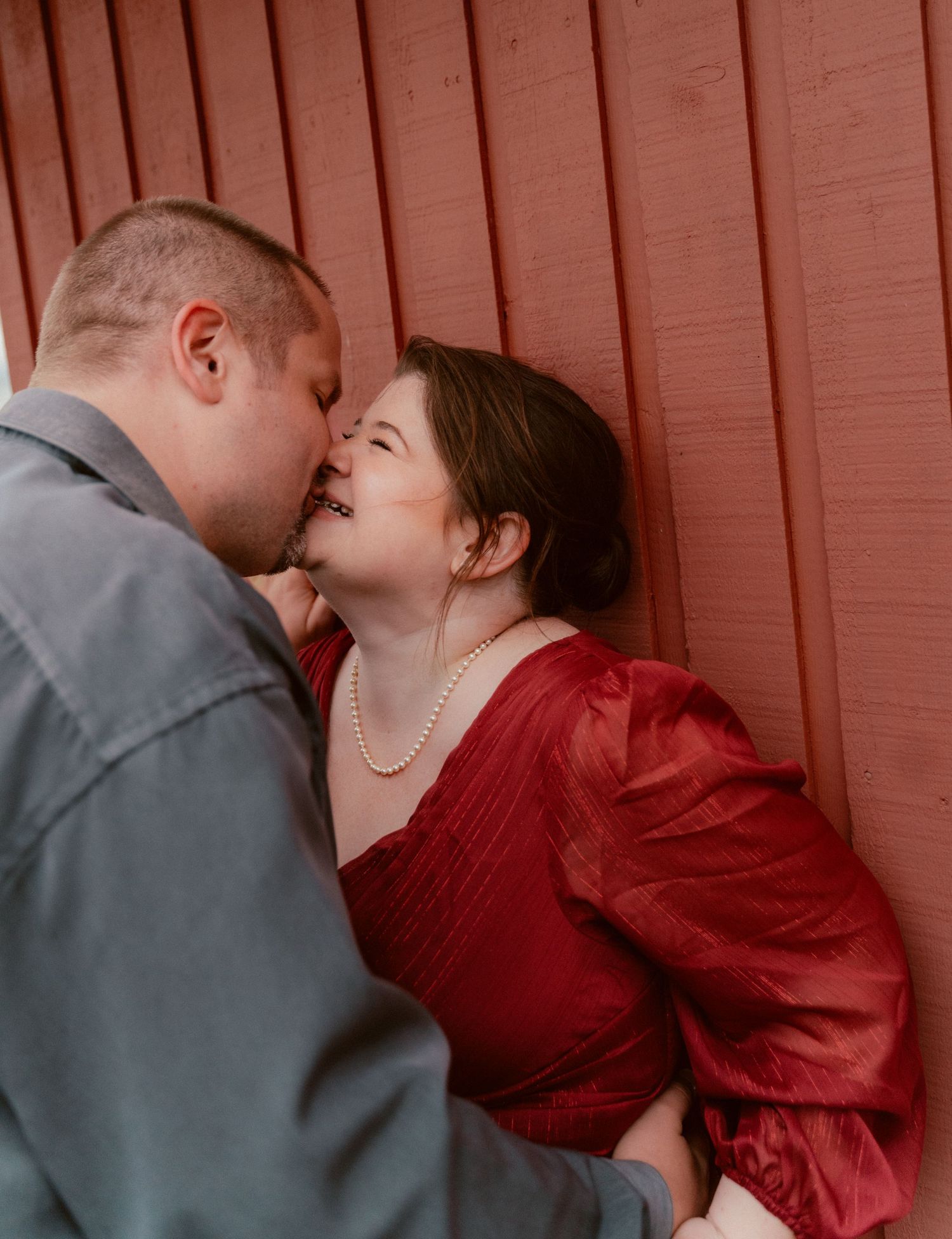 Tender moment between couple in red dress and gray suit against rustic red wall background.
