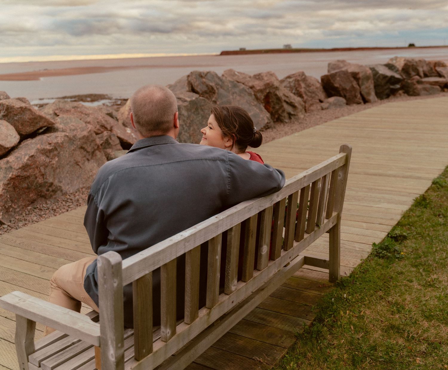 A couple sits together on a wooden bench overlooking a rocky beach during an overcast day.