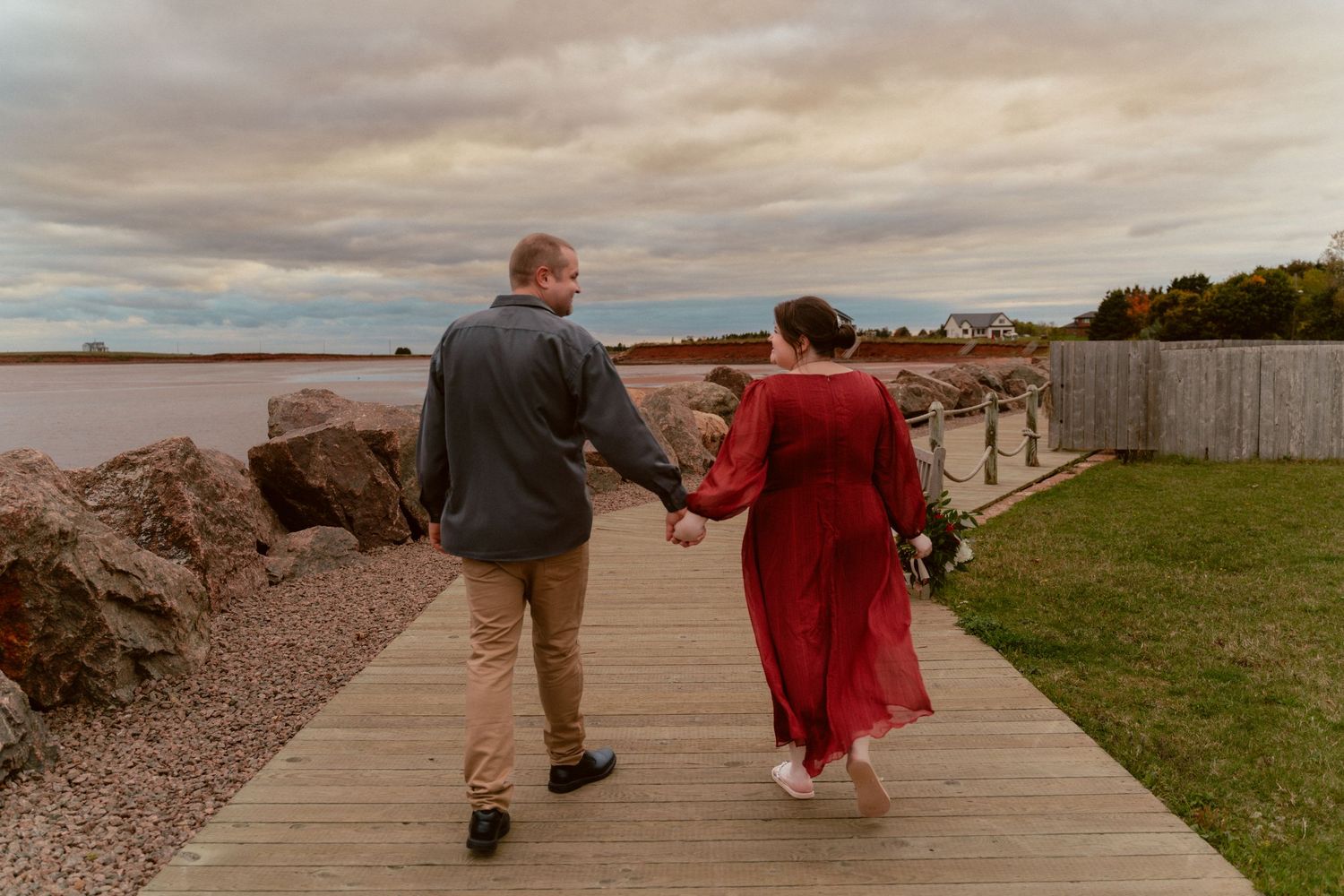 A couple walks hand in hand on a wooden boardwalk at sunset wearing formal attire.