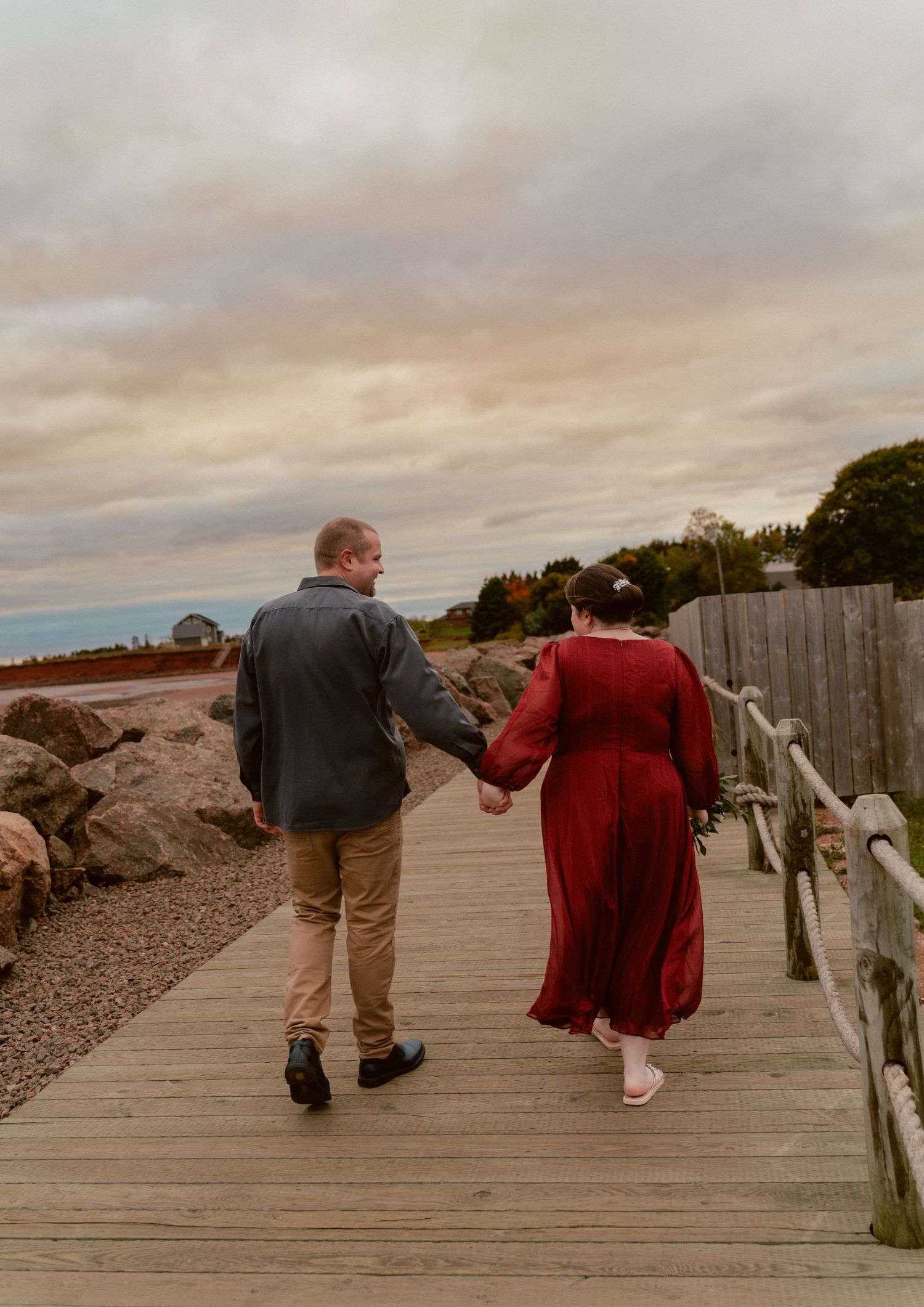 A couple walks hand in hand on a wooden boardwalk at sunset.