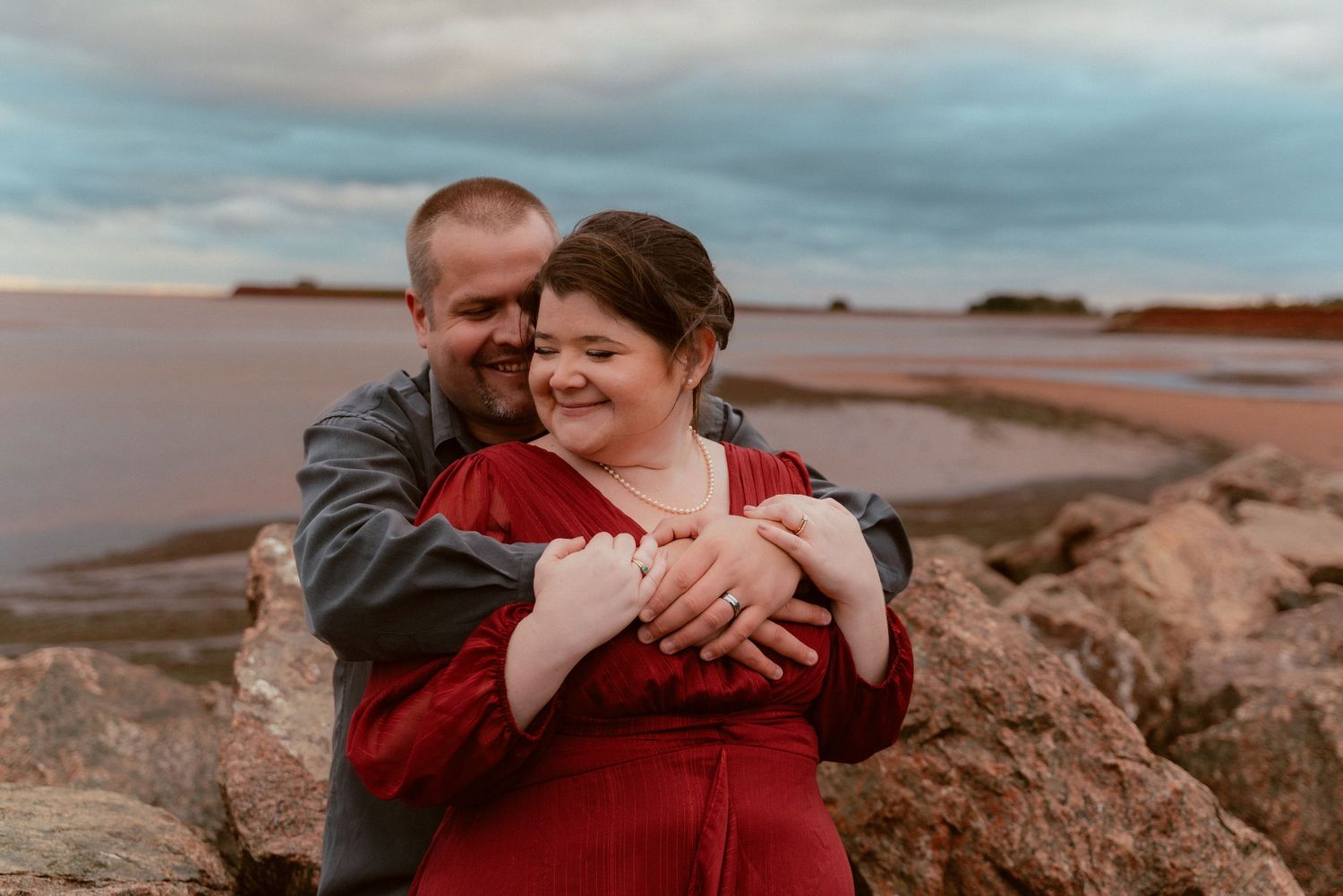 Romantic couple embracing on rocky shore during sunset with water in background.