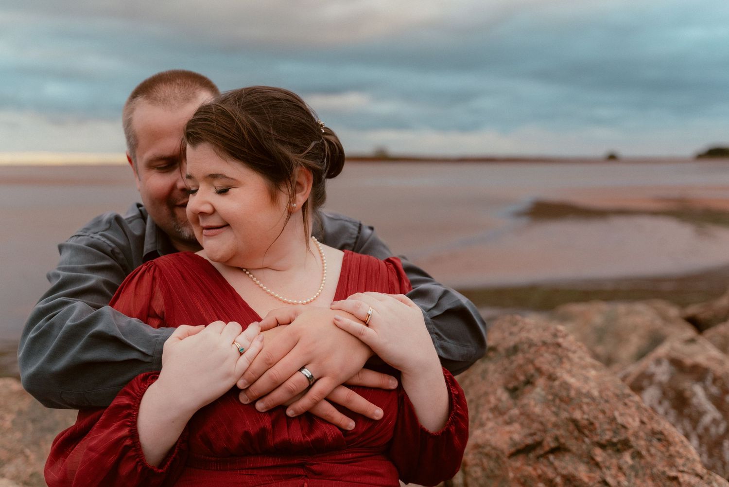 A couple embraces on rocky shore during sunset with dramatic cloudy sky.