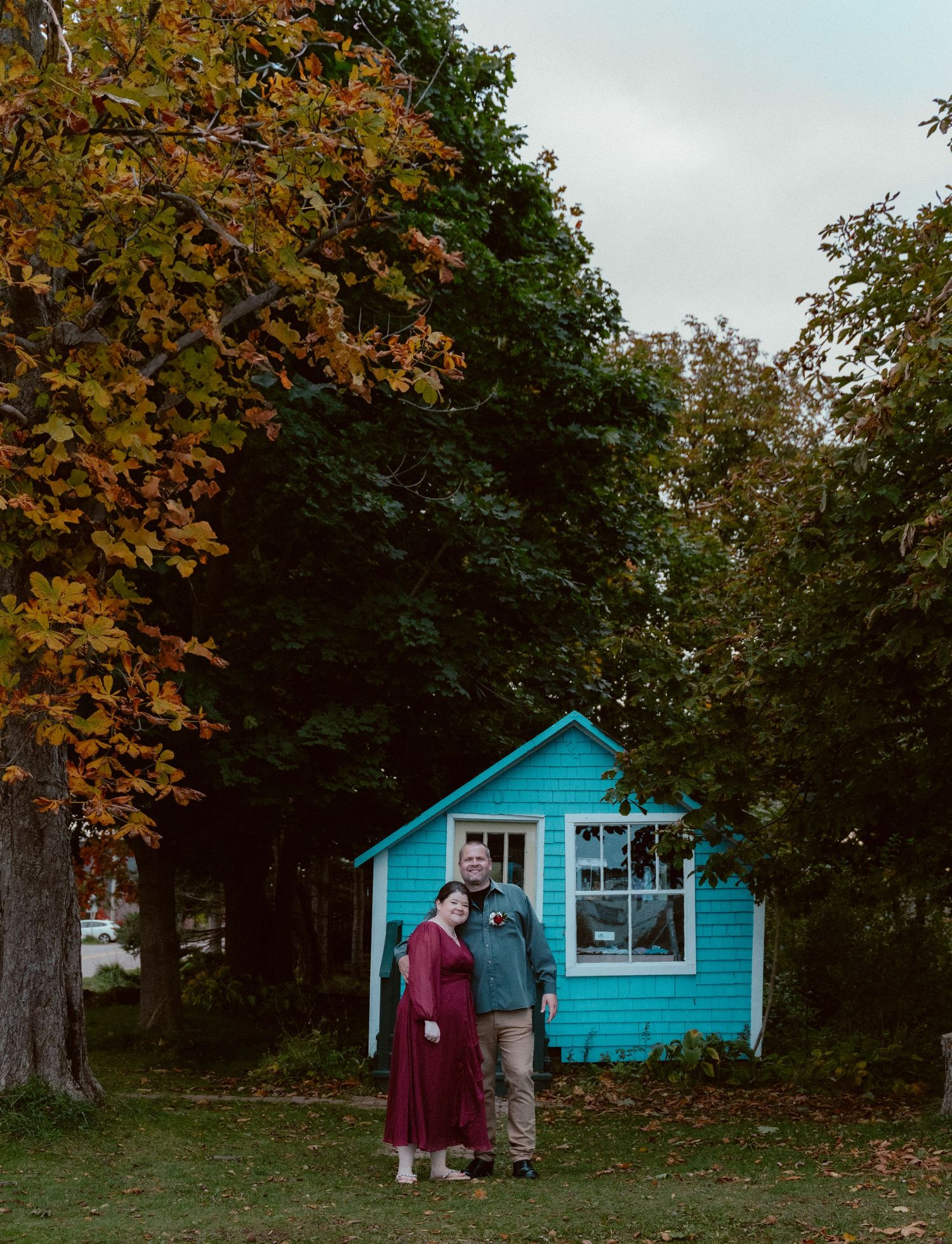 A couple stands together in front of a bright turquoise tiny house surrounded by fall foliage.