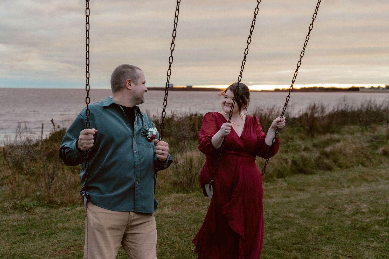 A couple shares a playful moment on swings at sunset by the water.