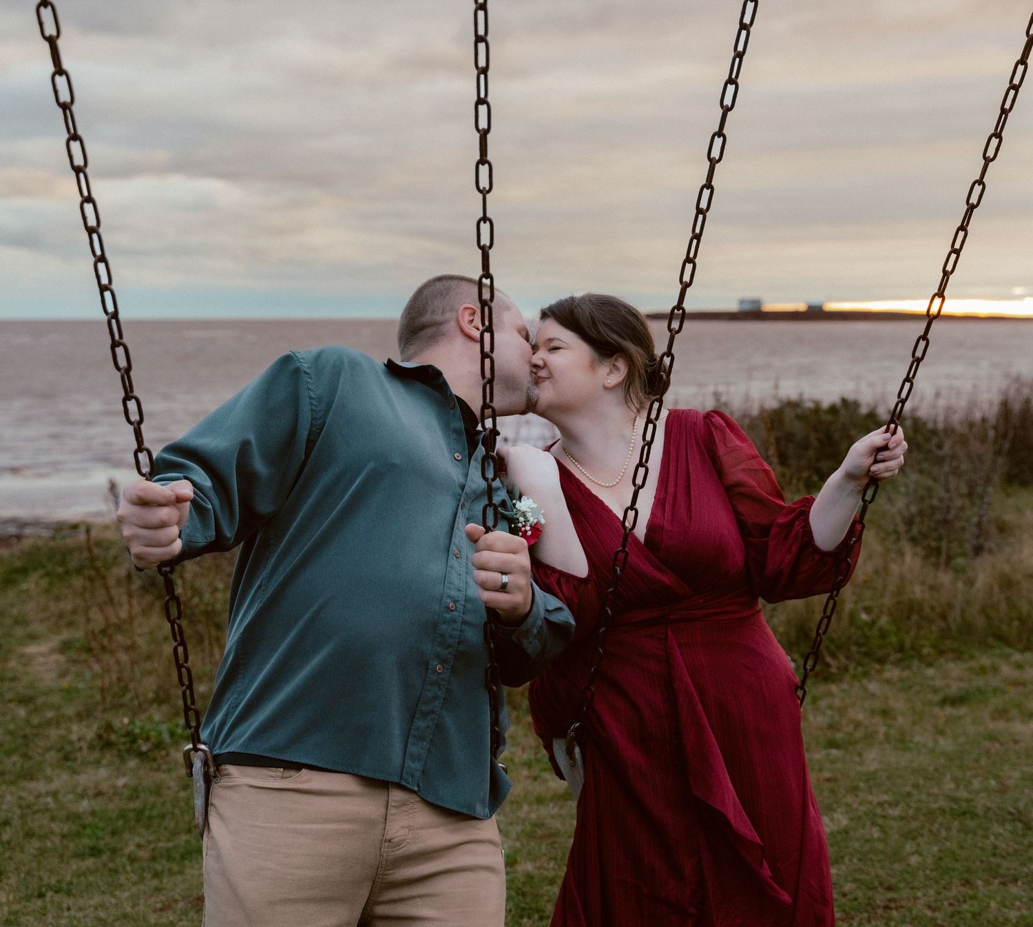 A joyful couple sharing a kiss while sitting on swings at sunset by the ocean.