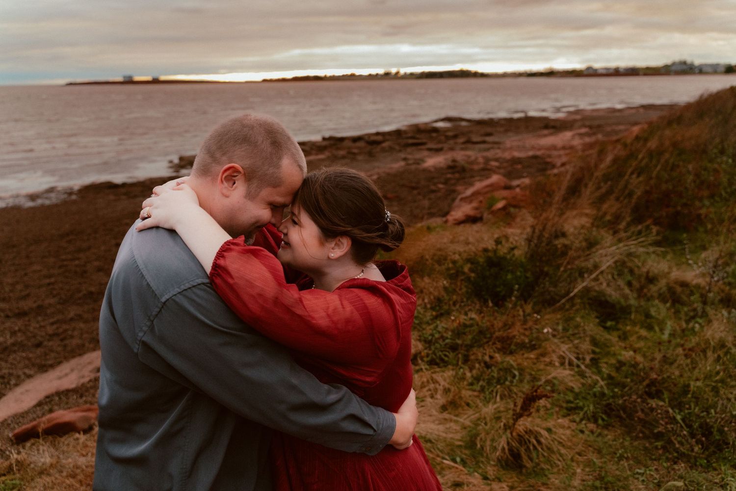 A romantic embrace on a rocky coastline at sunset with grey skies and ocean in the background.