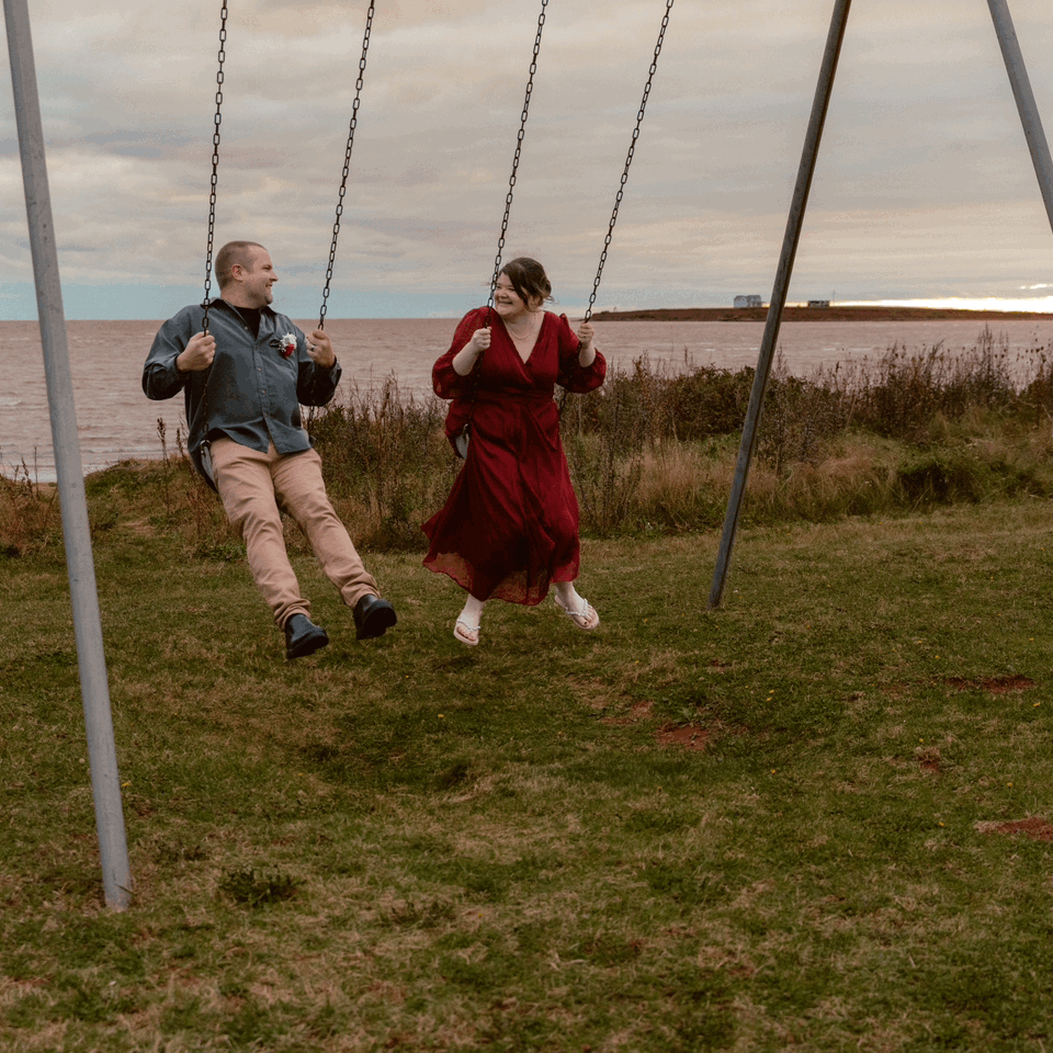 Two people enjoying swings at sunset near a waterfront, with one wearing a red dress and another in casual attire.