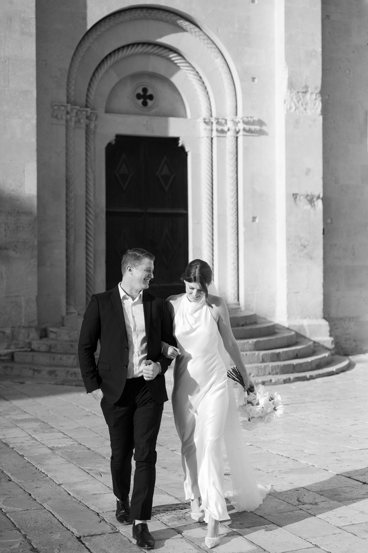 A couple exits a church after their wedding ceremony, with the bride in a flowing white dress and groom in black attire.