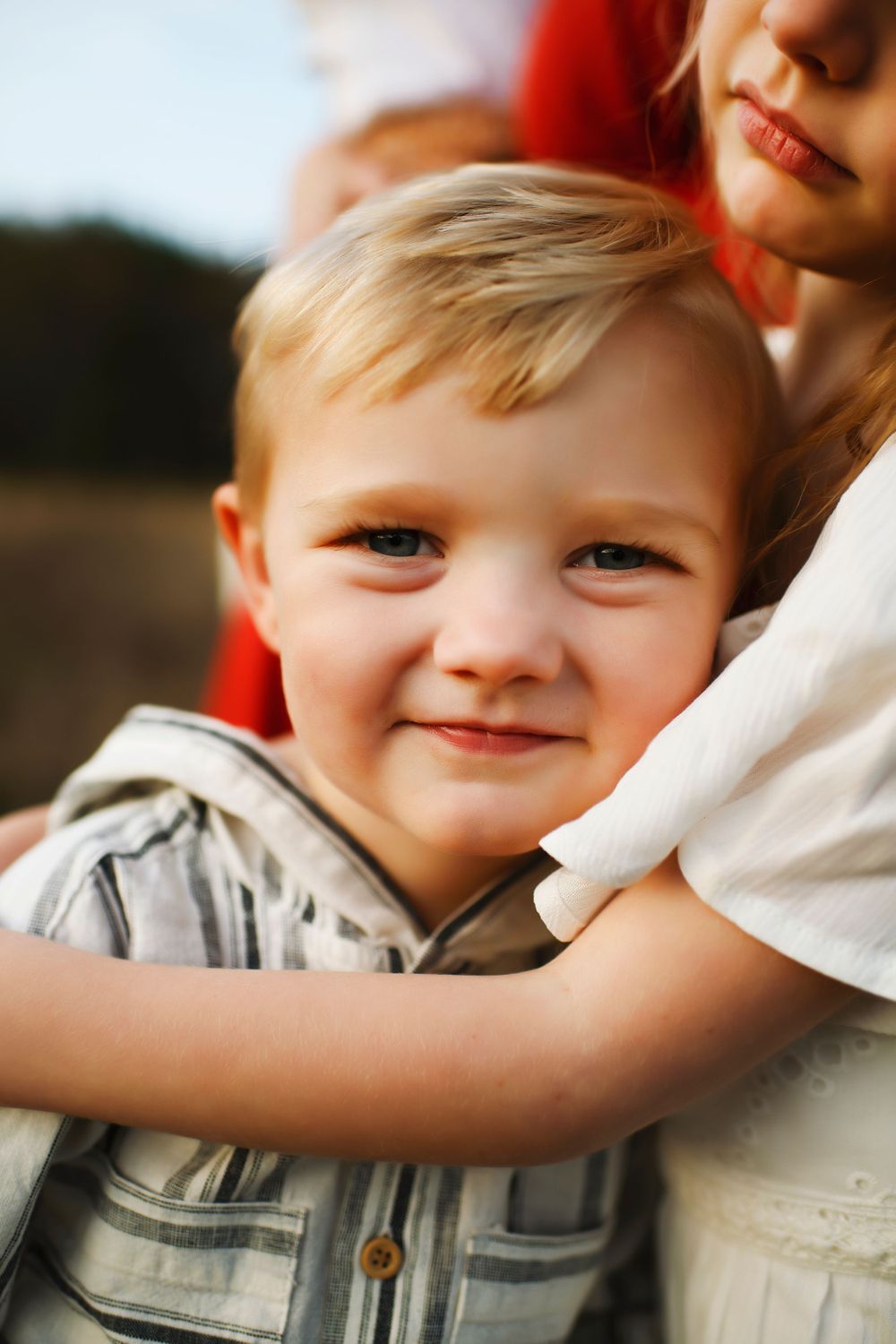 A close-up portrait in natural light showing siblings embracing against a blurred outdoor background.