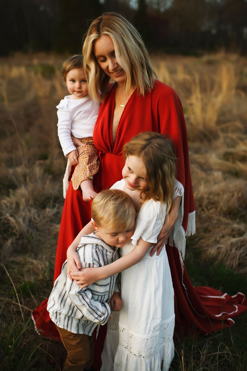 A woman in a flowing red dress stands with three children in a golden autumn field at sunset.