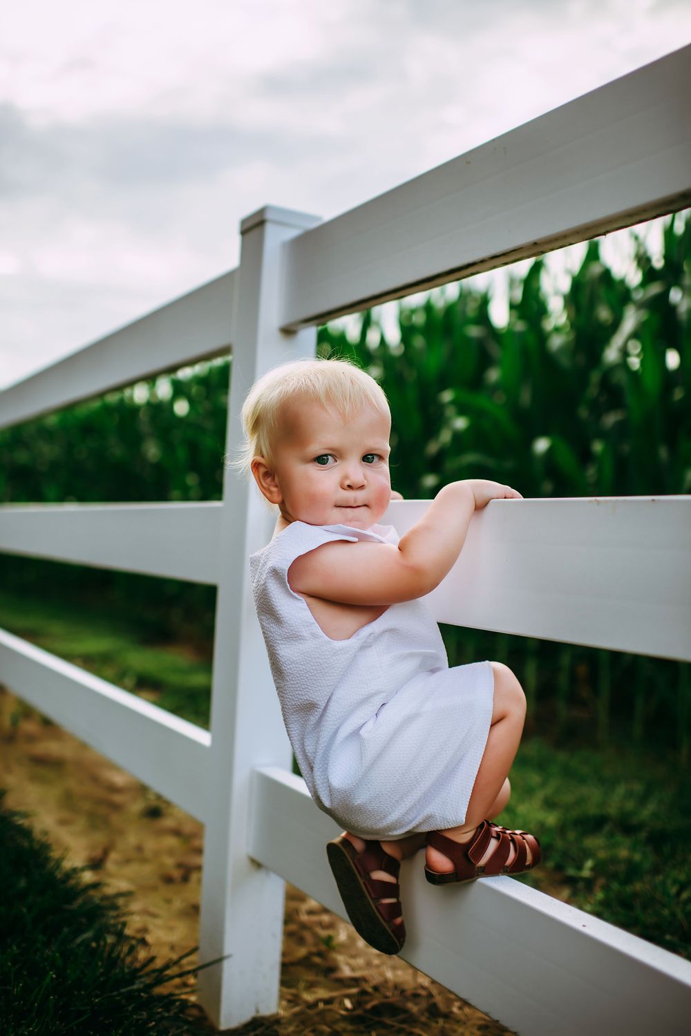 A small toddler in a white outfit and sandals climbs on a white fence rail while looking back at the camera.