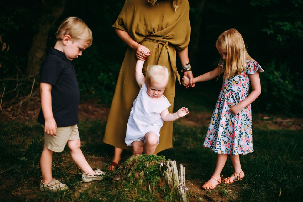Three children explore a forest path with an accompanying adult in a mustard-colored dress on a summer afternoon.