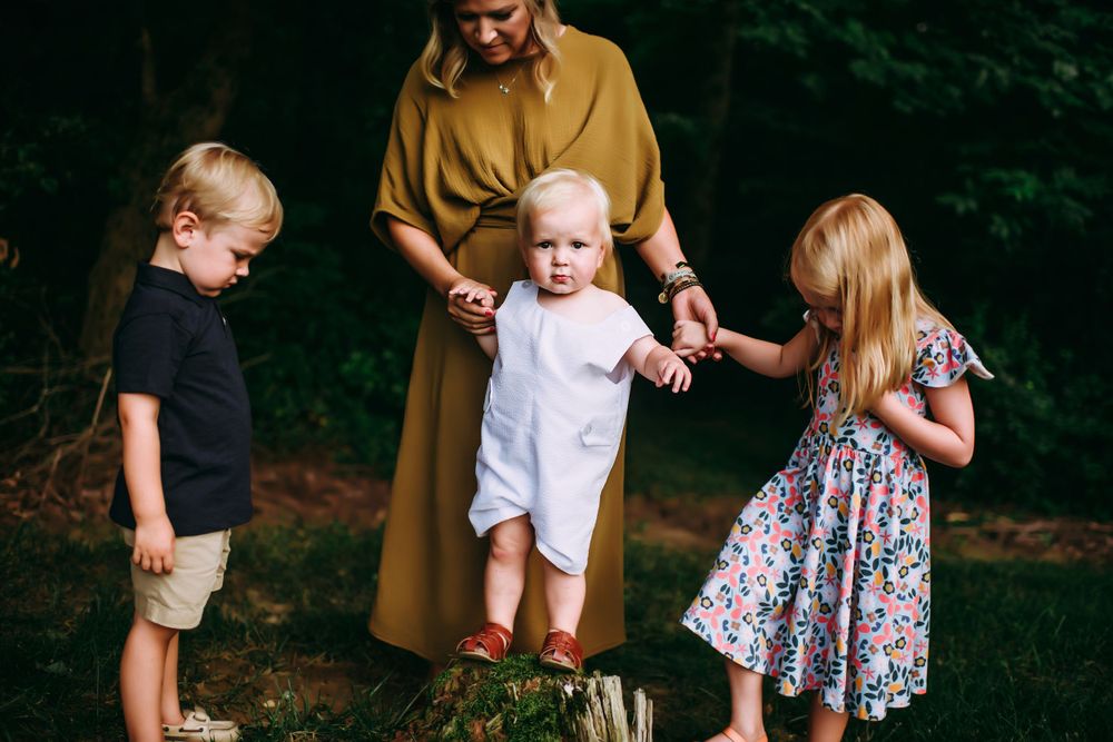 Children playing together outdoors in a wooded area with an adult in a mustard-colored dress.