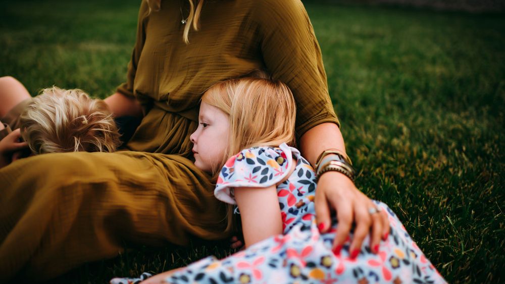 A young child in a floral dress relaxes on grassy ground with someone wearing brown clothing nearby.
