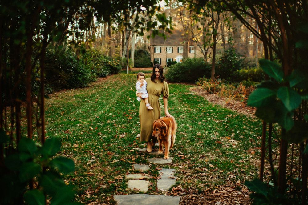 People walking with a dog down a tree-lined autumn path covered in fallen leaves.