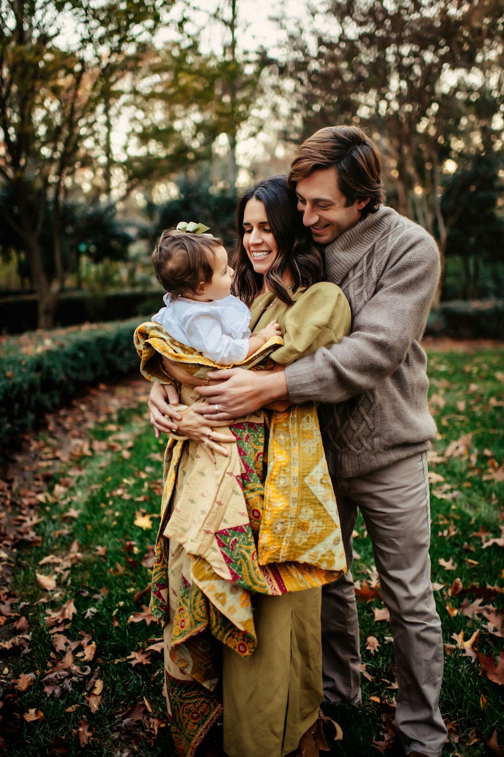 A family embraces while standing in a garden with fall leaves on the ground at sunset.
