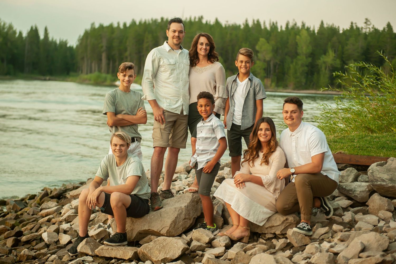A group dressed in casual light-colored clothing poses together on rocky riverbank with forest in background.