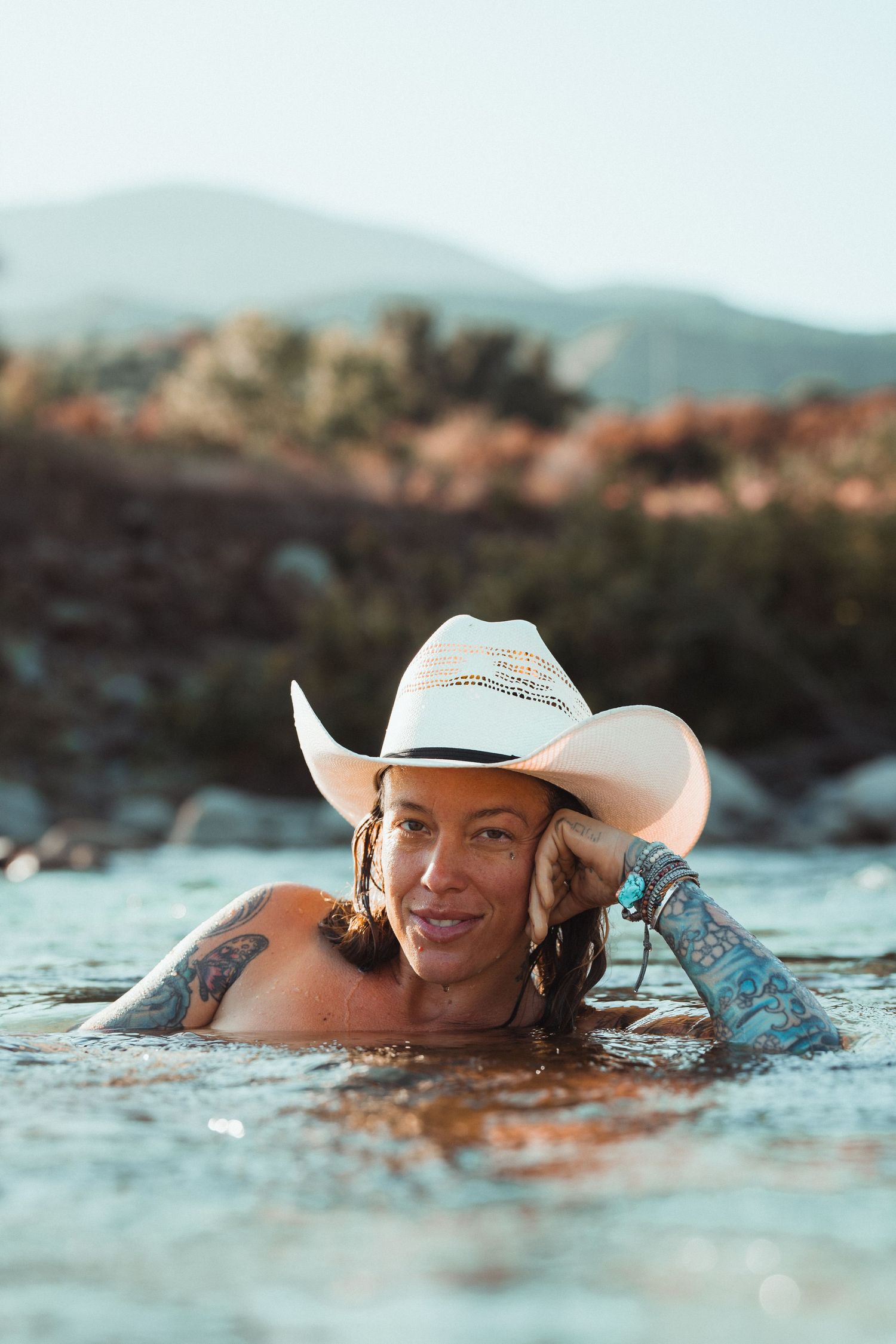 Person wearing white cowboy hat swimming in turquoise water with tattoos visible.