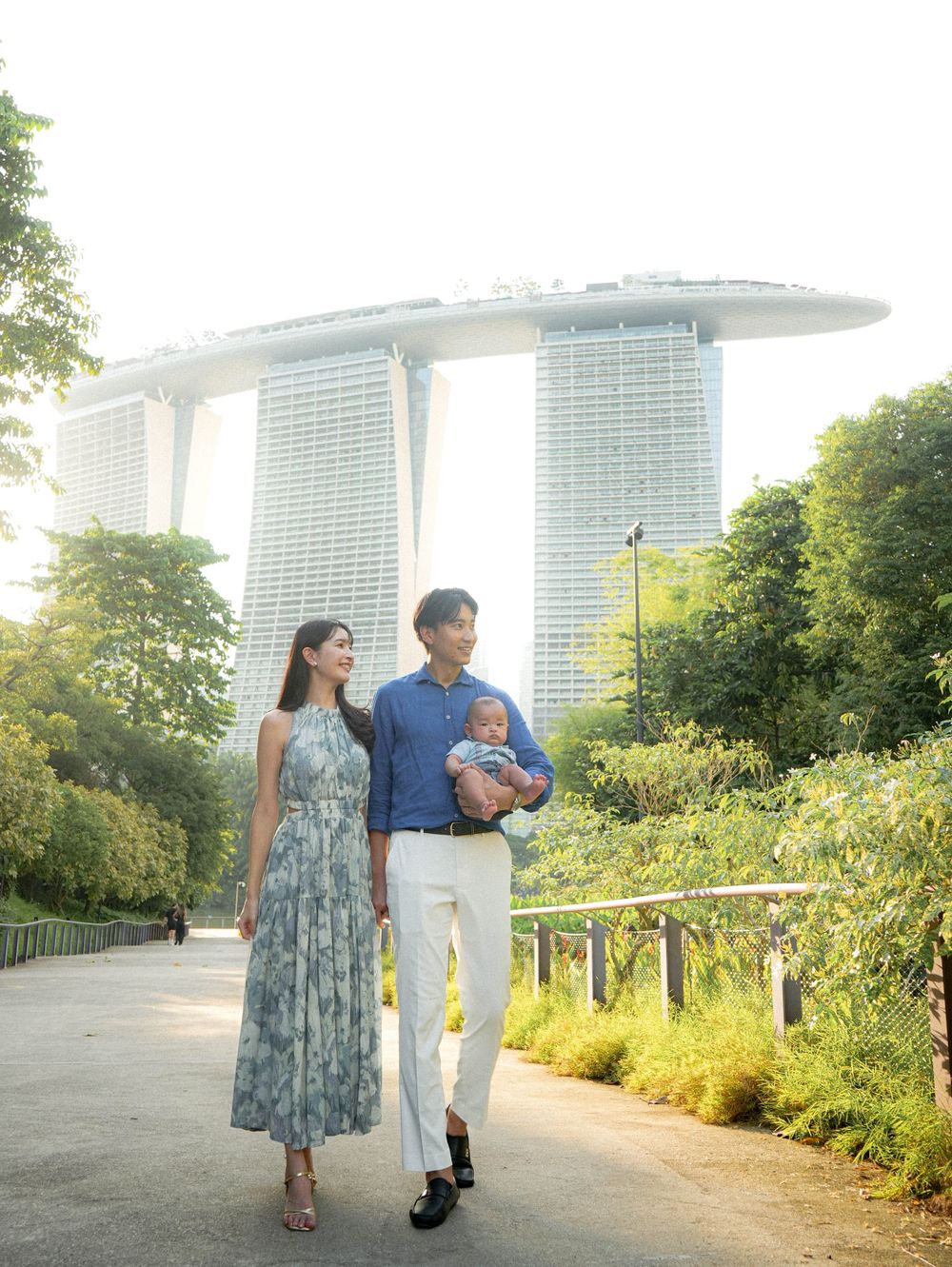 A couple and baby stroll along a garden path with Marina Bay Sands hotel in Singapore visible in the background.