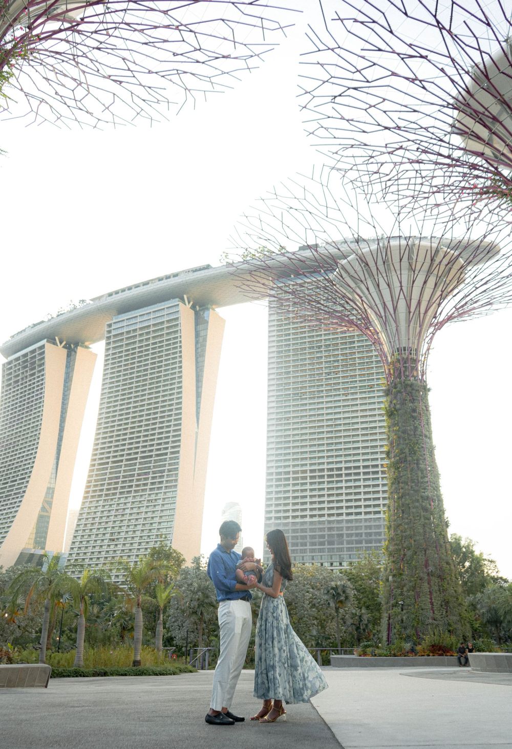 Romantic couple pose beneath Supertrees at Gardens by the Bay with Marina Bay Sands in background.