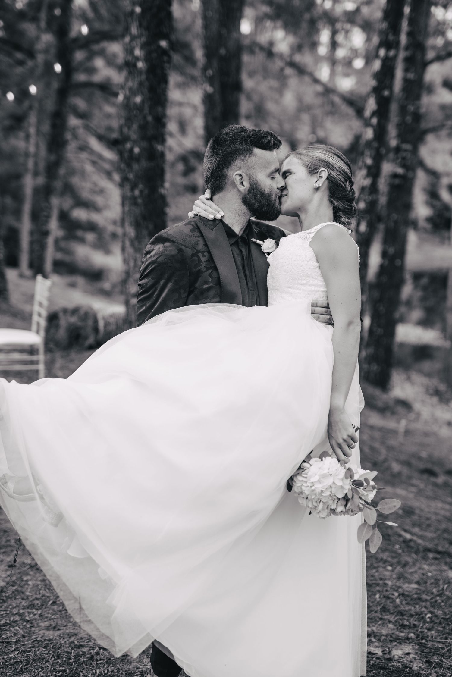 A romantic black and white wedding photo of a couple kissing in the woods while the bride's dress flows in the wind.