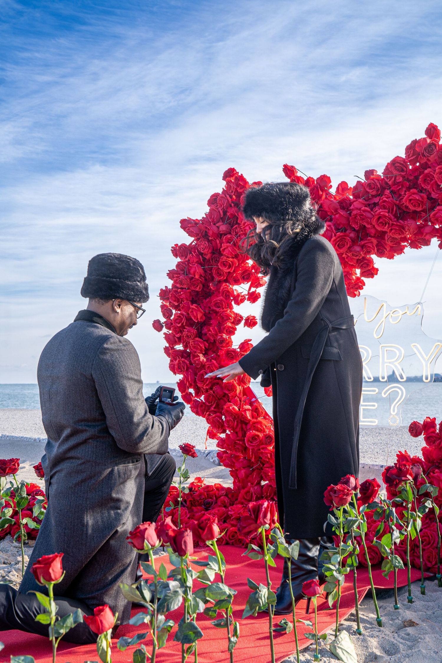 A romantic beach proposal setup with a red heart-shaped balloon arch and rose petals on a red carpet by the ocean.