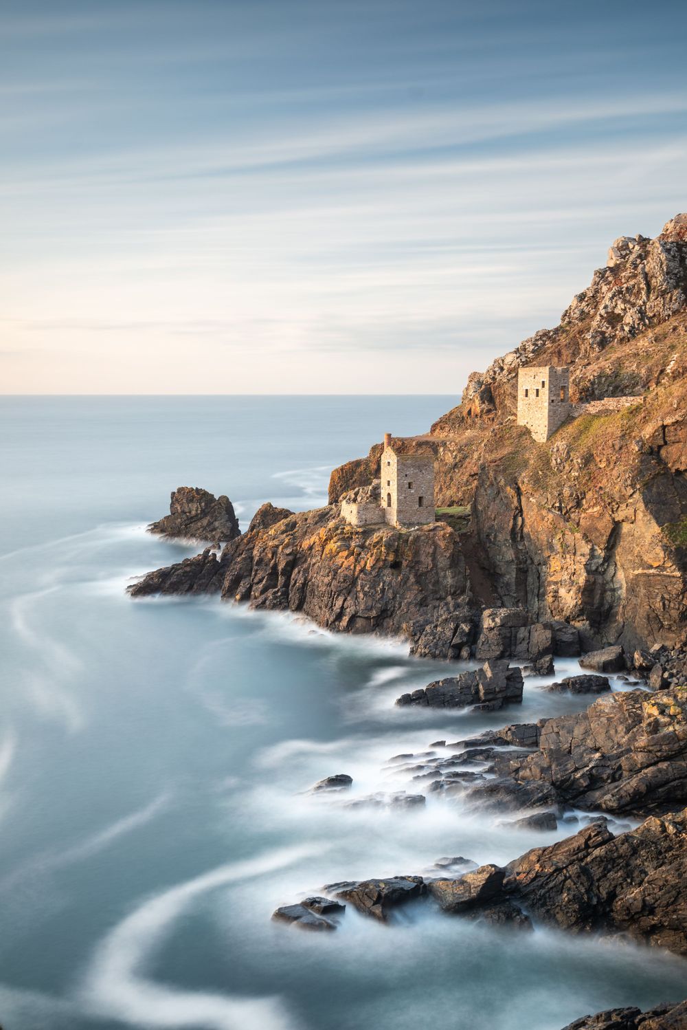 Historic stone buildings perched on rugged cliffs above misty waves crashing on Cornwall's dramatic rocky coastline.