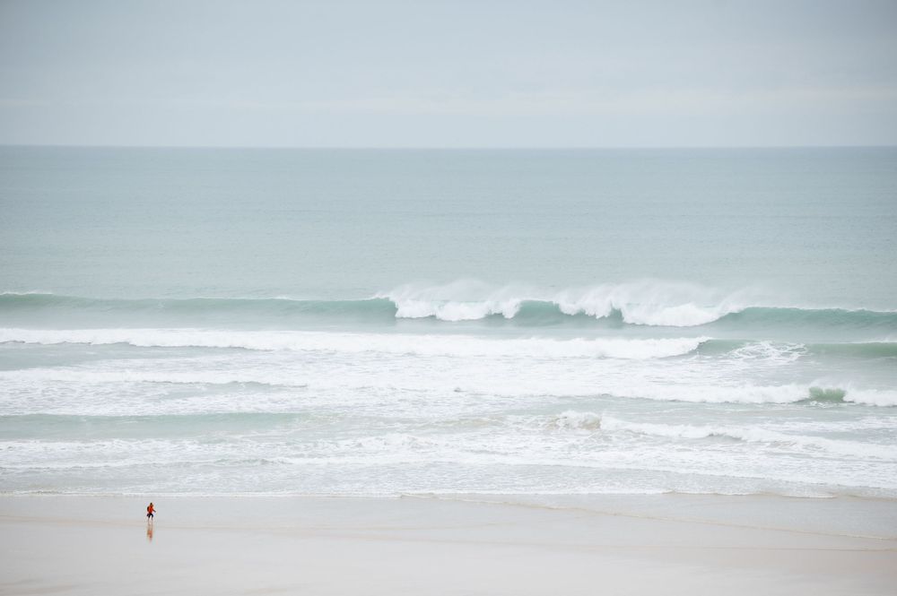A lone person walks along a misty beach with rolling waves and overcast sky in the distance.