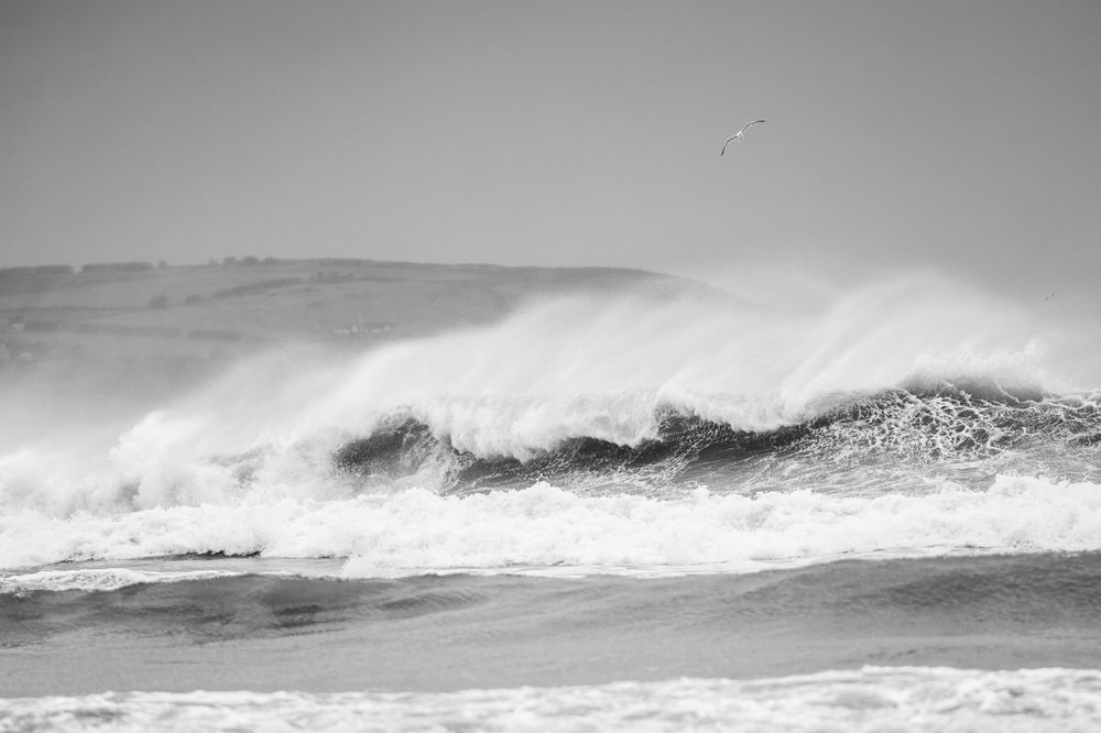 A powerful ocean wave crashes and sprays white foam against a stormy gray sky in this black and white seascape.