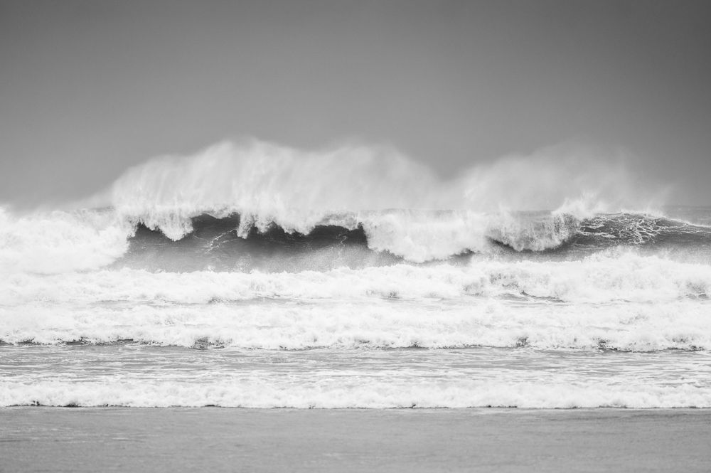 Large ocean wave crashes with dramatic white spray against a gray cloudy sky in black and white photography.