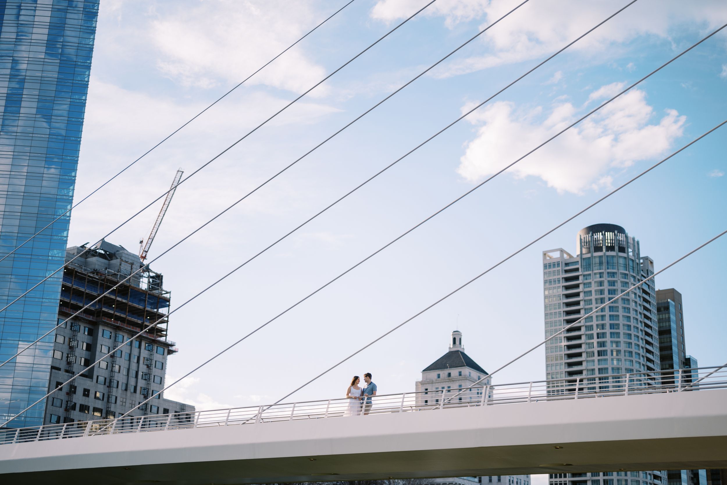Silhouetted figures stand on the white curved bridge against a blue sky with downtown Milwaukee buildings in background.