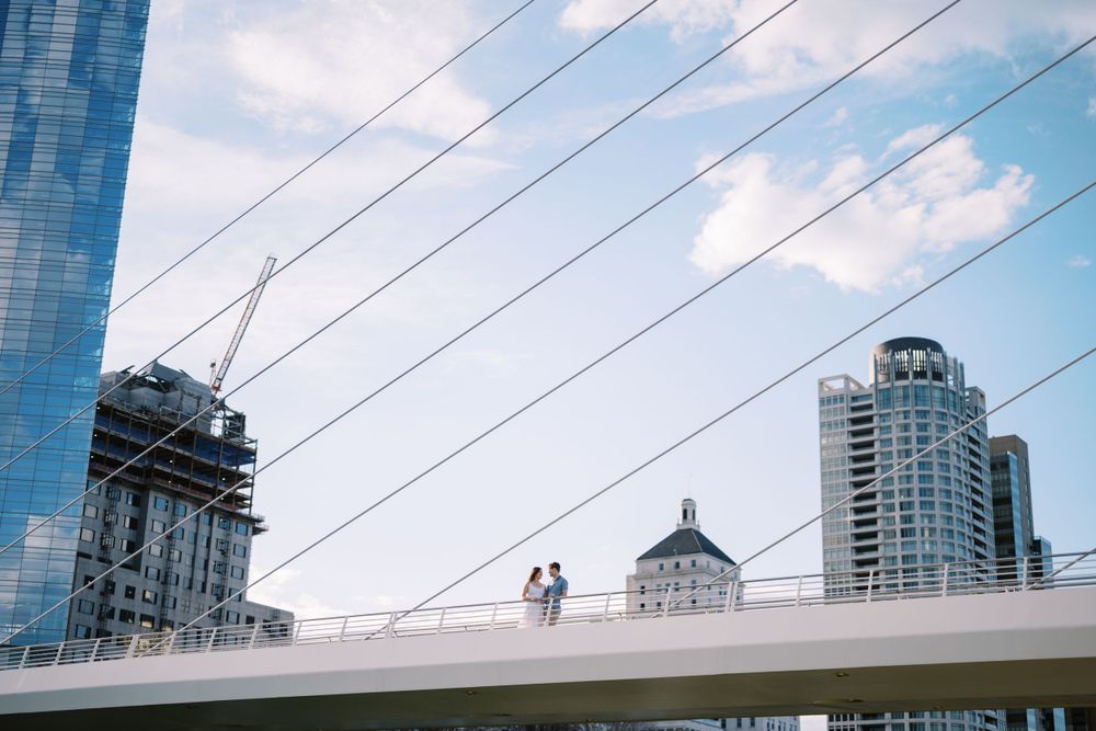 Silhouetted figures stand on the white curved bridge against a blue sky with downtown Milwaukee buildings in background.