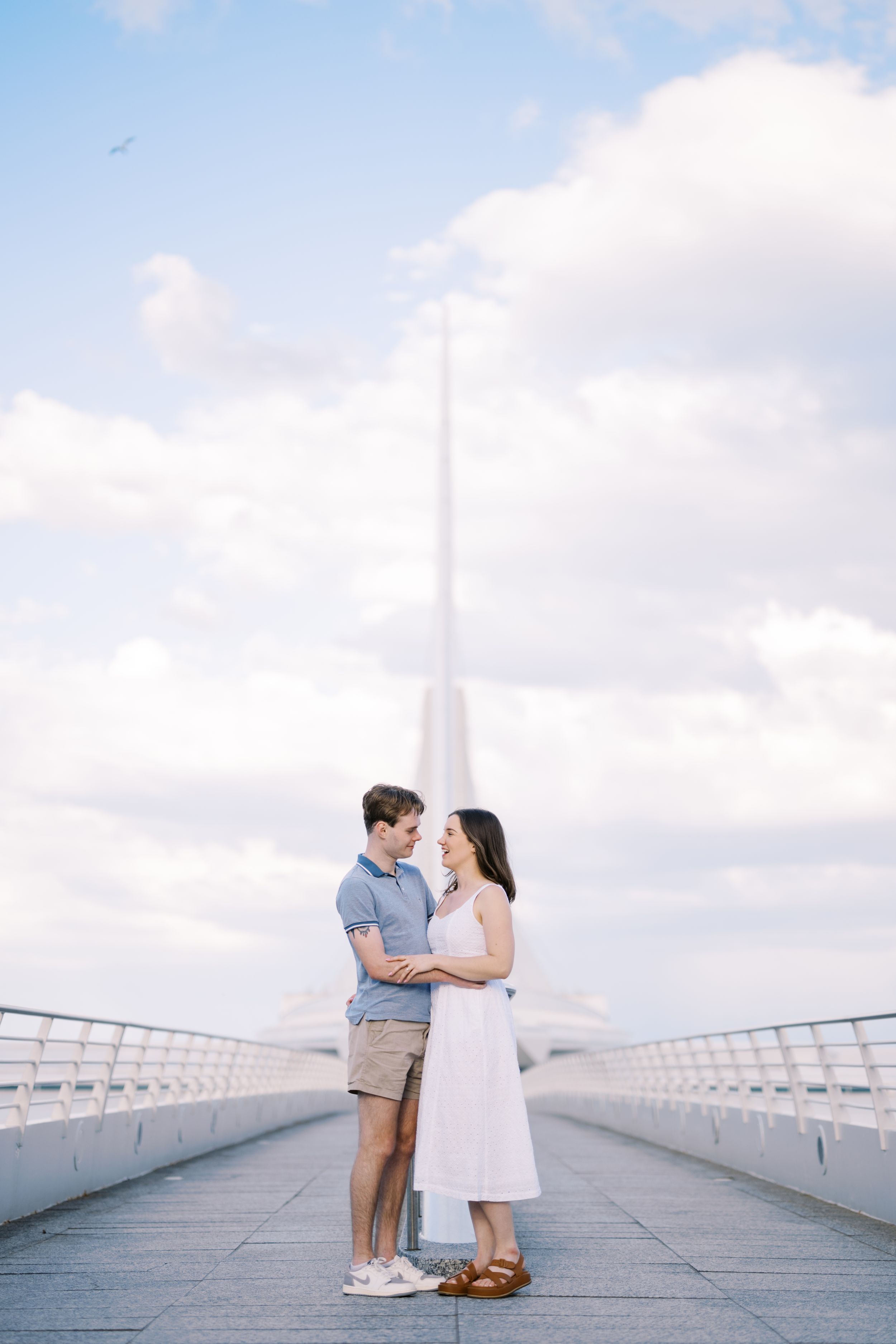 A couple embraces on a bridge walkway with soft clouds and architecture in the background.