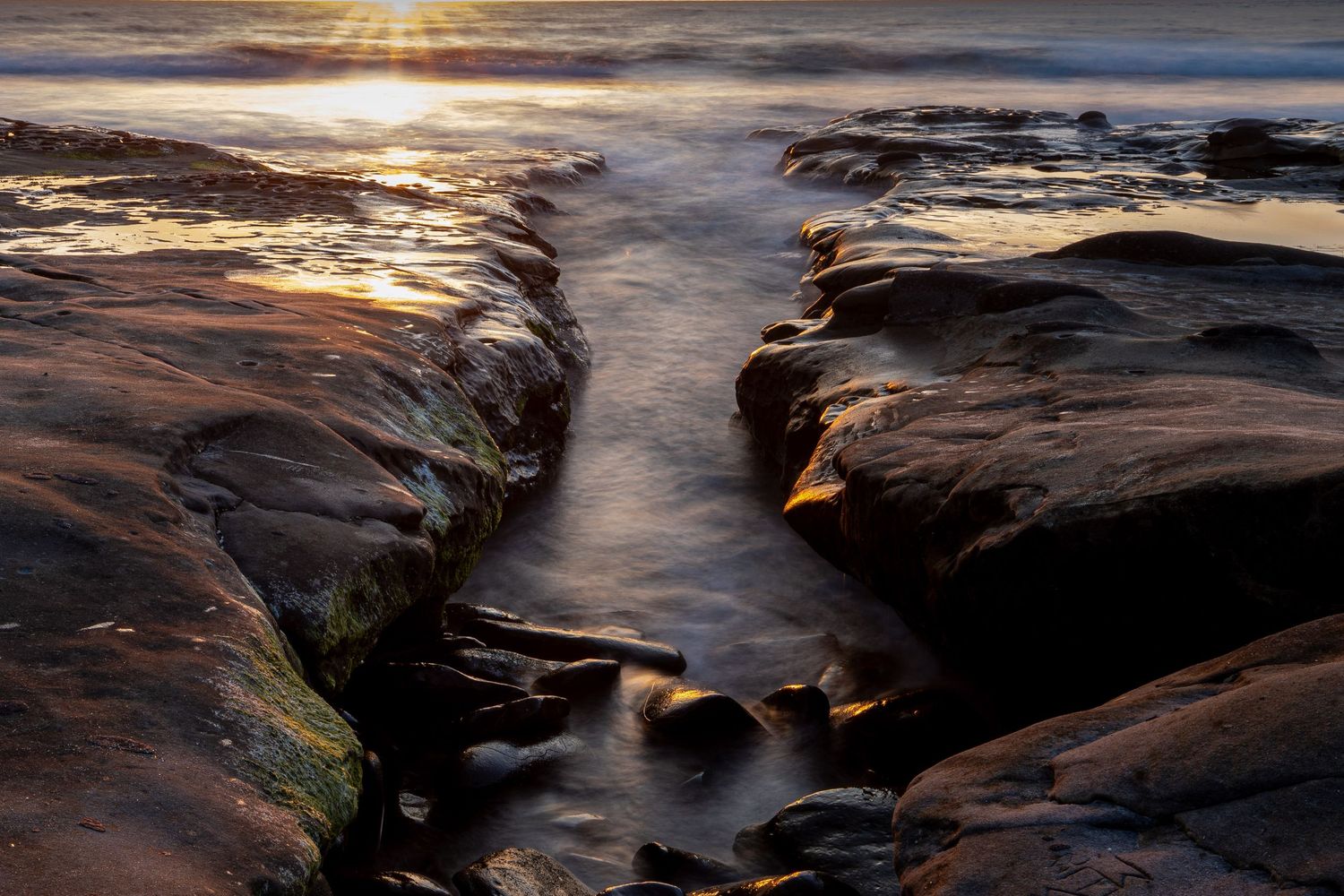 Dramatic sunset over rocky coastal terrain with water flowing through a narrow channel between cliffs in La Jolla, California.