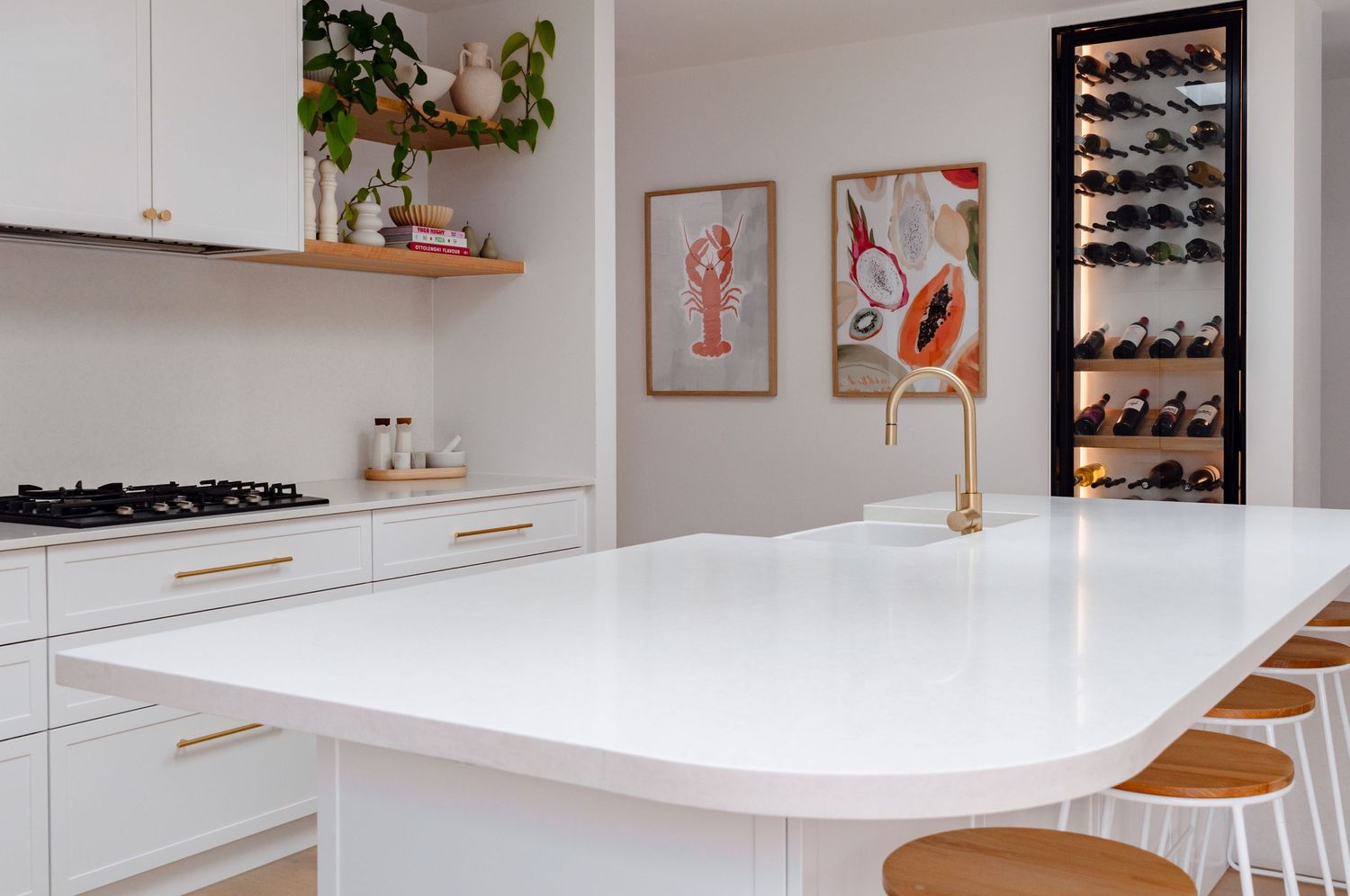 Modern white kitchen with marble countertop, wooden bar stools, wall art prints, and a built-in shoe storage cabinet.