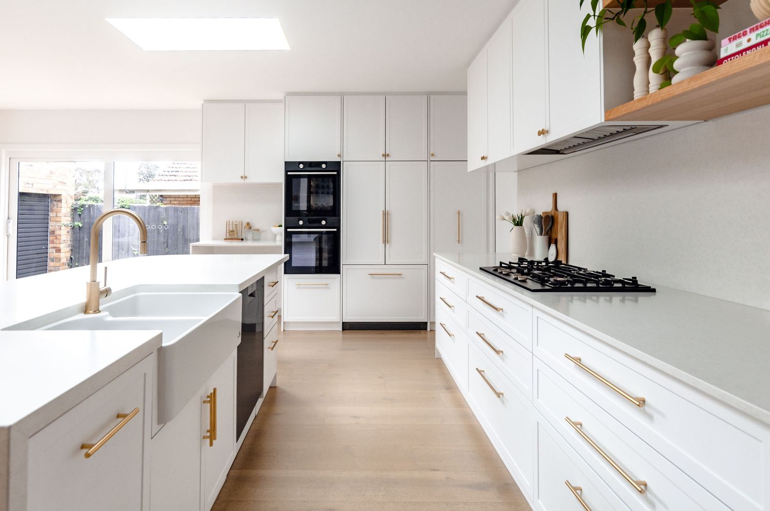 Modern white kitchen with marble countertops, gold hardware, wooden shelving and light hardwood floors.