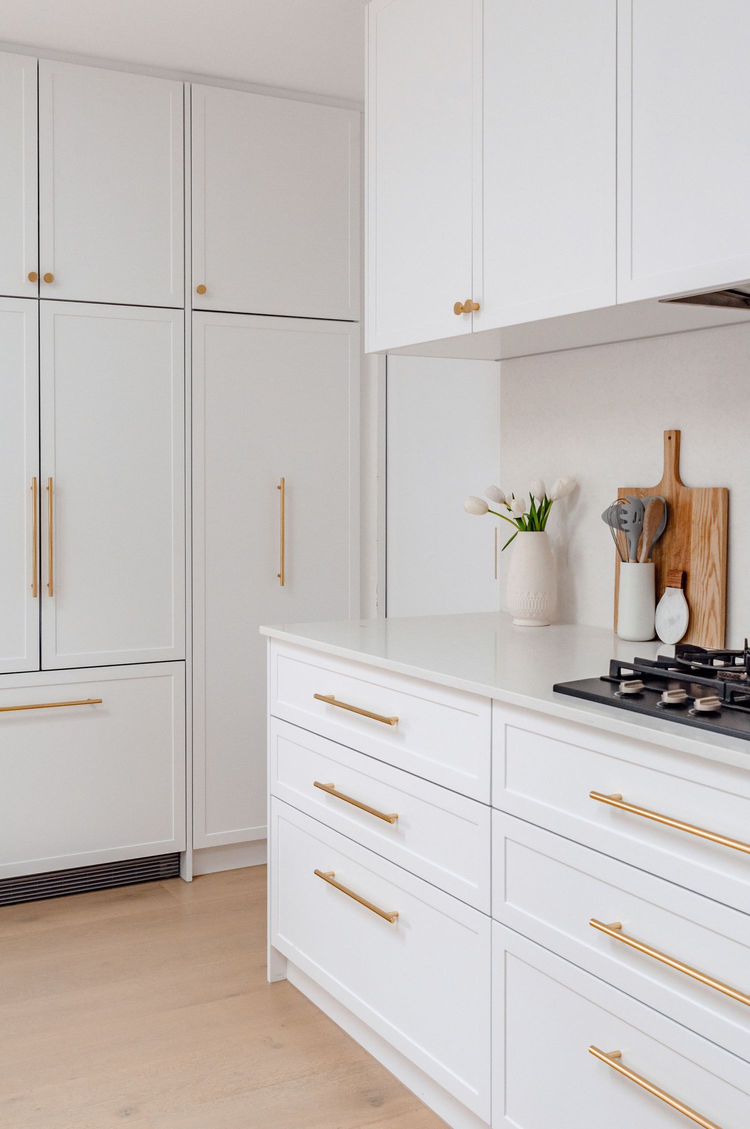 Modern white kitchen with tall cabinets featuring brass hardware, white countertops and light wood flooring.