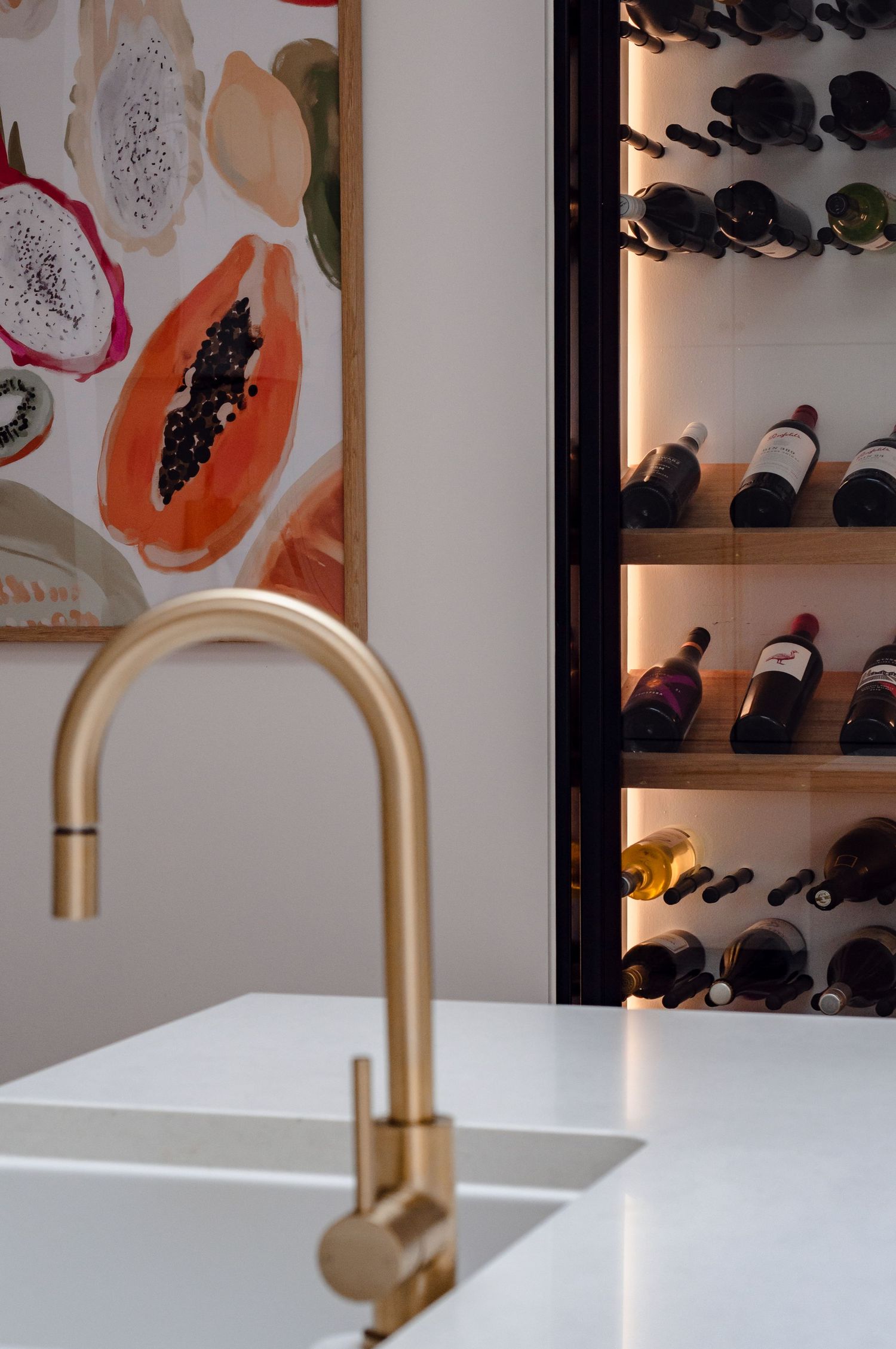 Close-up of gold faucet against white countertop with abstract fruit artwork and illuminated wine storage in background.