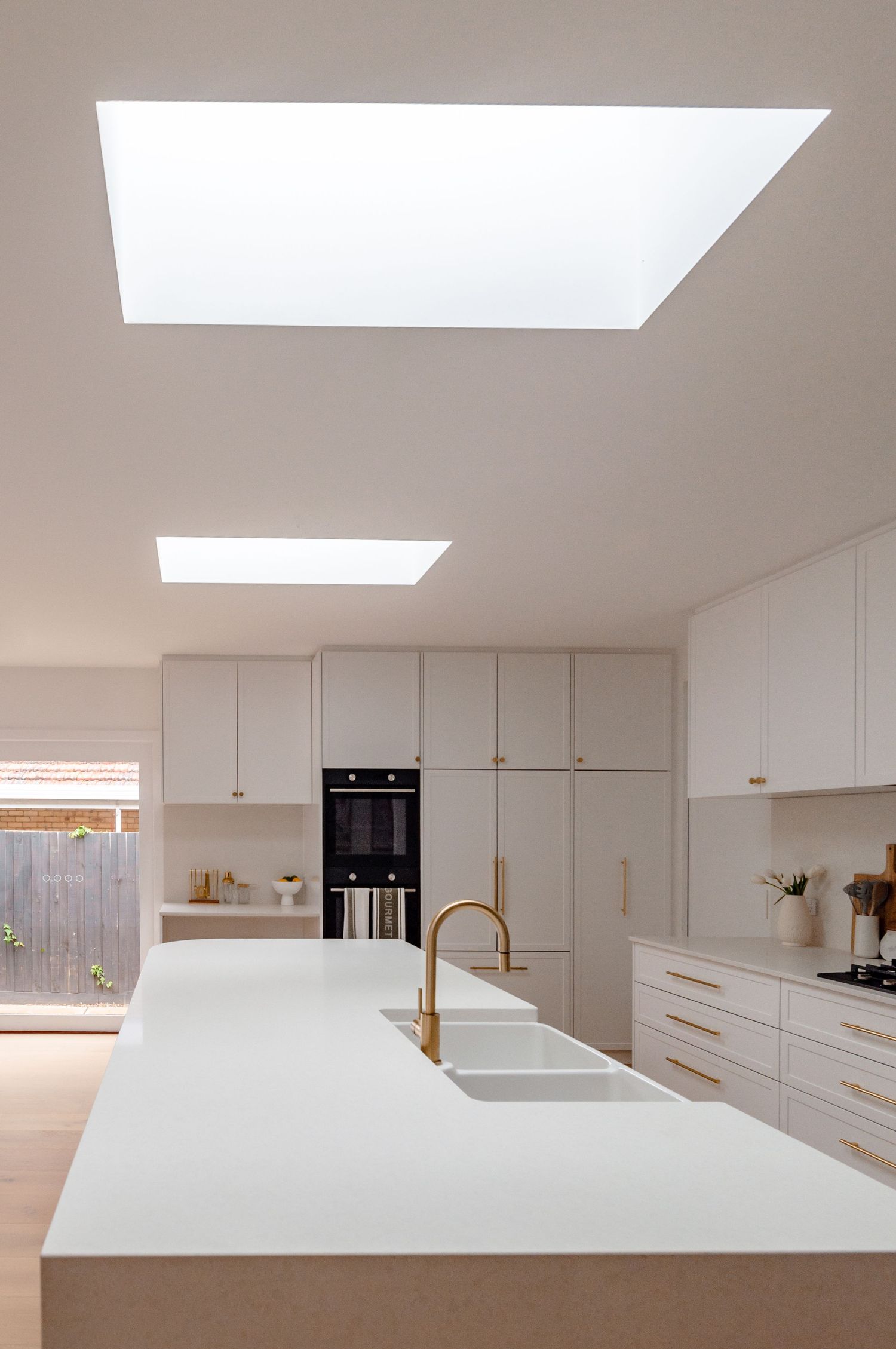 Contemporary white kitchen featuring skylights, built-in appliances, and a minimalist island with gold faucet.
