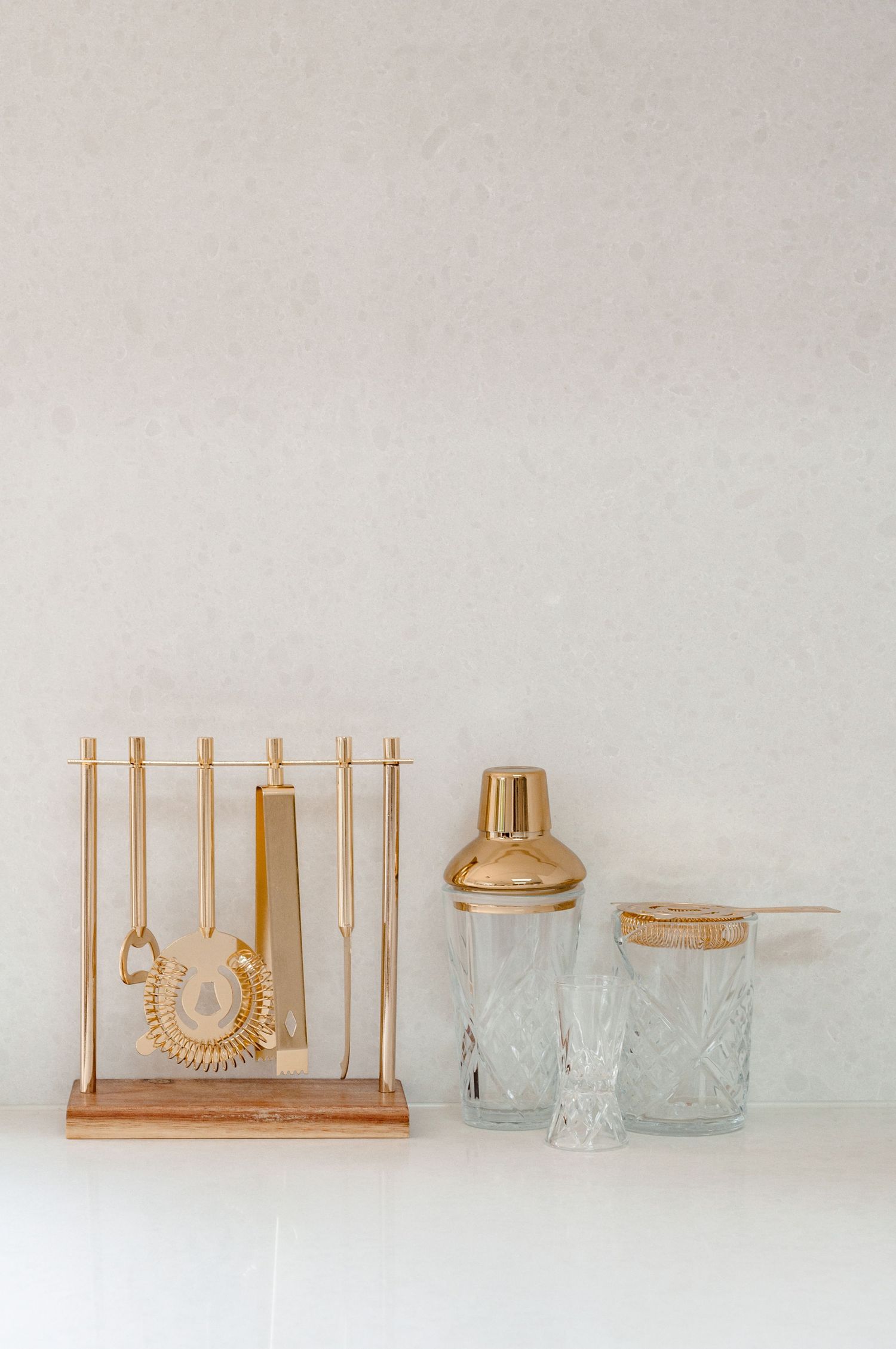 Gold bar tools and cocktail shaker set with textured glass tumblers on white counter against neutral wall.