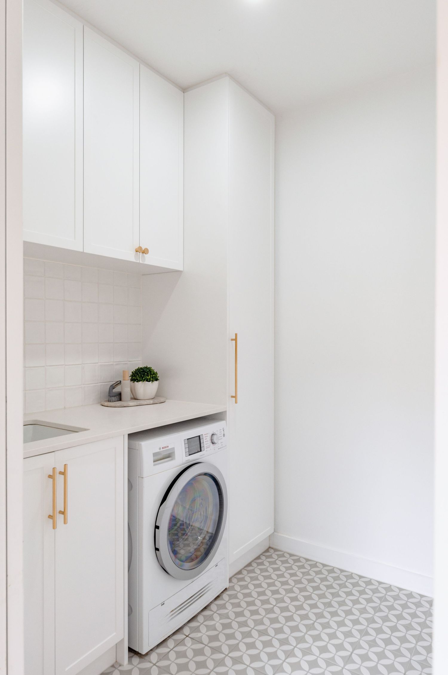 Modern minimalist laundry room with white cabinets, gold hardware, and patterned floor tiles features built-in washer unit.
