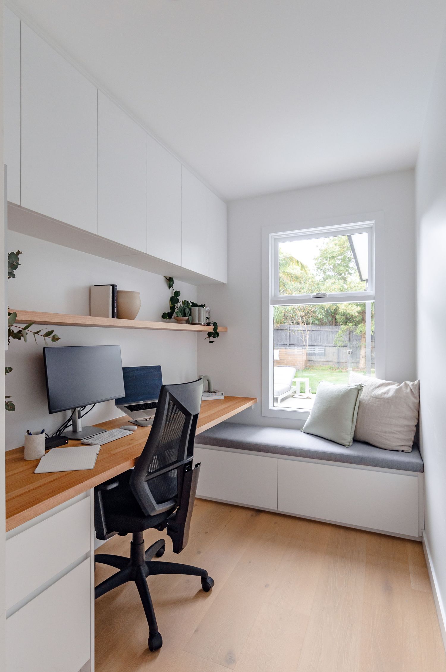 Modern home office with built-in desk, white cabinets, floating shelves and cozy window seat with grey cushions.