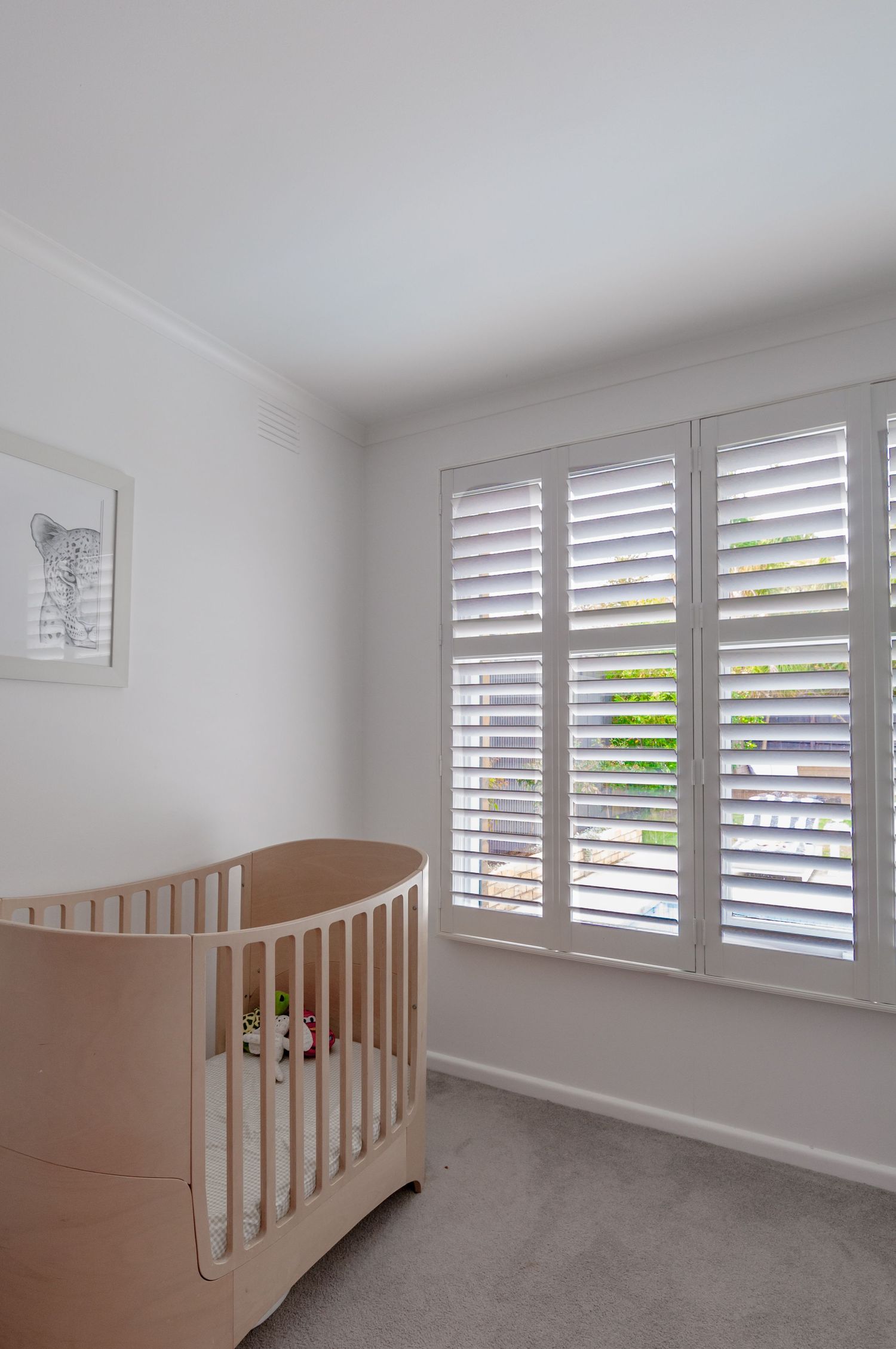 Modern nursery room with white plantation shutters, a light wooden crib, and neutral grey carpeting.