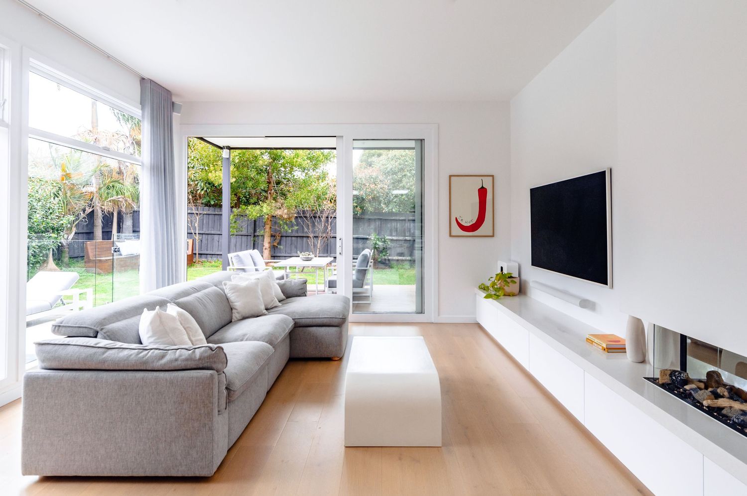 Modern minimalist living room with gray sectional sofa, white ottoman and floor-to-ceiling windows overlooking a garden.