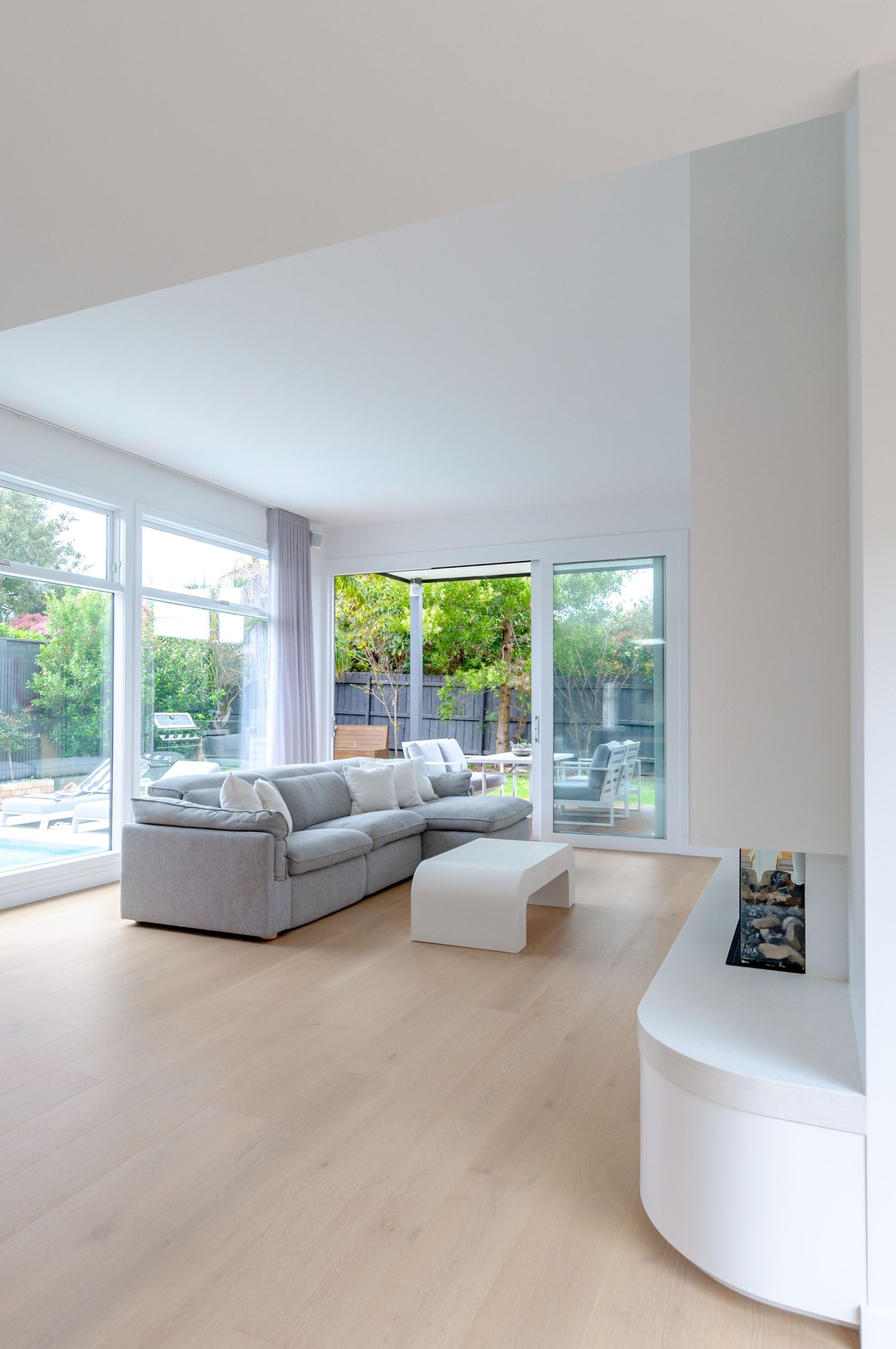 Modern living room with light wood floors, gray sectional sofa, and large windows overlooking a garden patio area.