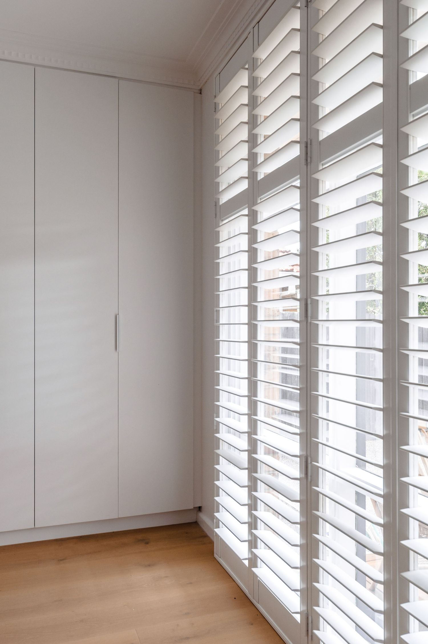 Bright corner room with white plantation shutters and built-in wardrobes featuring hardwood flooring.