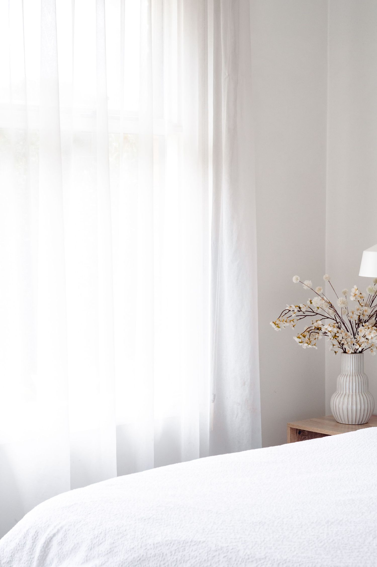 Minimal bedroom interior with white bedding, sheer curtains, and dried flowers in a ribbed vase on side table.